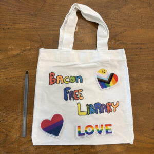 a mini tote bag next to a pen for size reference with rainbow wording spelling out Bacon Free Library, an iron on bisexual pride flag heart, an iron on pride flag heart, and an iron on of the word love in a rainbow pattern